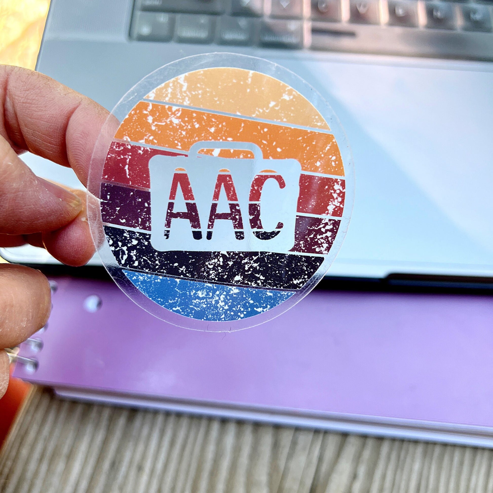 Clear circle AAC speaker with retro colors.  There is an AAC device cutout with AAC written inside.  An SLP is holding the AAC decal in front of a computer.