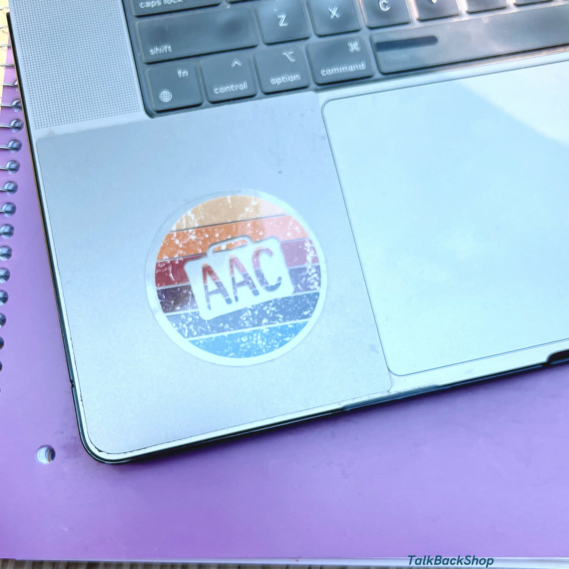 Clear circle AAC speaker with retro colors.  There is an AAC device cutout with AAC written inside.  The AAC decal is on a laptop.